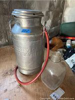 Photo showing the full metal milk can with red rubber tube attached and clear glass jug placed beside it on the floor, environment indicates storage or basement area.