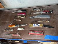 Overview of fourteen pocket knives of various styles and sizes laid out on a table showing soiling and rust.