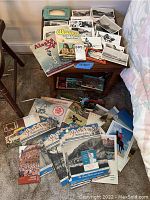 Wide view showing assortment of vintage postcards, pamphlets and ephemera on floor and table, mainly Alaskan travel items and Pearl Harbor story piece