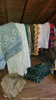 Vintage bedding items draped over and near a metal clothes rack, including multiple blankets of various patterns and colors such as gray floral geometric, white fringed, and colorful zigzag.