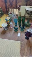 Photo of assorted vintage colored glassware including vases, bottles, candlesticks, jars, and a blue and white urn, laid out on a wooden floor with yellow fabric background.