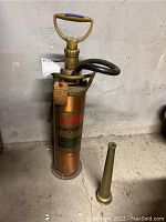 Full view of vintage copper-colored metal fire extinguisher standing upright with black rubber hose attached and a detached metal hose nozzle placed on the floor beside it.