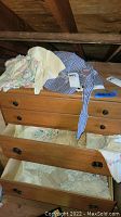 Pile of vintage linens and folded fabrics on a wooden dresser with some drawers slightly open showing more linens inside.