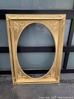 Full view of large rectangular gold spray painted wooden frame with ornate floral and scroll decorations and an oval opening in the center.