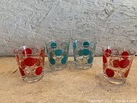 Four mid century modern style drinking glasses on a beige surface against a textured wall, two with red dots and two with blue dots, all with concentric circle pattern.