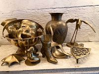 Wide view showing all brass items grouped together including the vase, masks, duck, bird, boot, umbrella, trivet, and scoop.
