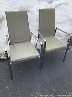 Front view of two Calligaris Italian armchairs with beige upholstery and chrome frames set outdoors on pavement with snow in the background. Visible scratches on upholstery.