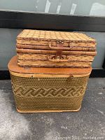 Two vintage picnic baskets stacked; larger woven basket with patterned design on bottom, smaller rectangular woven wicker basket with leather latch on top. Both show age wear; larger basket missing handle.
