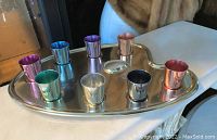 Nine colored metal shot glasses arranged on a metal artist's palette-shaped tray with recessed slots for each glass.