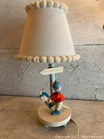 Full view of the vintage child's lamp showing the figurine, lamp base, and cream fabric lampshade trimmed with pom-poms.