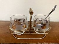 Front view of cream and sugar glass containers with floral motifs in a gold-toned metal holder with spoon included