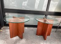 Pair of vintage side tables with round glass tops and teak cross bases shown outdoors, viewed from front