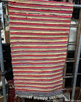 Full view of vintage rag rug hung vertically showing red, beige, and blue horizontal stripes and fringe at bottom.