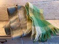Two wool textiles: one is a beige and cream shaded shawl folded, the other is a checkered green, yellow, and cream wool blanket with fringes.