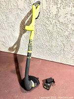 Full view of RYOBI 18v cordless string trimmer standing against a wall with charger on ground.