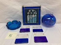 Full view of the lot showing the blue glass ashtray, four coasters, witches' ball suncatcher, and stained glass painting in a metal frame.
