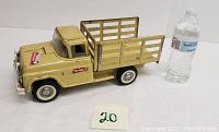 Side view of the yellow pressed steel Buddy L farm truck with wooden slat bed, next to a water bottle for size comparison showing about 14 inches length.
