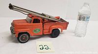 Side angle showing the orange metal truck with pile driver arm extended, next to a water bottle for size reference.