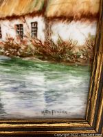 Close-up of the bottom corner of the framed oil painting showing the thatched roof home, water, and artist signature 'M.De Fraine'.
