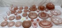 Wide view showing a large assortment of pink depression glass including cups, saucers, bowls, and plates with ornate patterns, all arranged on a white background.