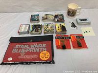 Photo showing all items together: multiple trading card stacks, mug, black and red folder of Star Wars blueprints, two pendant packages, and a single loose card.