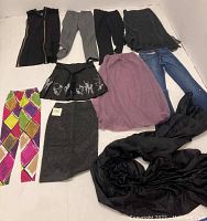 Photo showing full lot of clothing items arranged on the floor. Includes colorful Moschino pants, various skirts, denim jeans, black shawl, and a black sleeveless garment with zipper.
