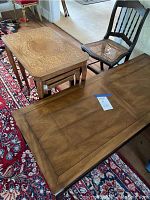 Photo showing wood coffee table with smooth top and two nesting tables with ornate design, and antique chair with broken caned seat in the background on red patterned rug.