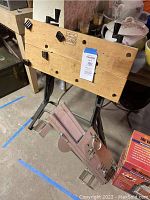 Wooden top of the folding saw table with black clamps and black metal legs visible, displayed with boxed propane heater on floor.
