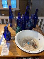View of nine cobalt blue glass bottles, one clear glass bottle, green glass insulator, white metal basin, and wooden cutting board on table near window.