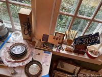 Overall view of vintage plates, glassware, key, fan, license plate and other items arranged on windowsill and nearby surfaces