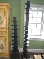 Two tall black metal shelves with multiple tiered platforms, standing side by side near a window and decorative wooden cabinet.
