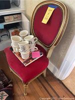 Red velvet oval-back wooden chair with gold trim, set of four white ceramic mugs with black classic composer illustrations stacked on the chair seat, atop a hardcover book.