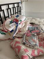 Photo showing several folded handmade quilts on a bed including floral and geometric designs, plus clear plastic bags with smaller fabric pieces.