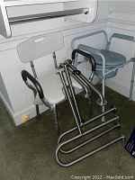 Photo showing a white plastic shower chair with backrest, light grey raised toilet seat with padded armrests, and three metal grab bars with black grip handles.