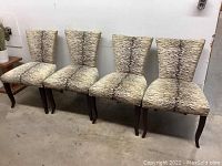 Four zebra print upholstered dining chairs with wooden cabriole style legs, arranged in a row against a white wall and concrete floor.