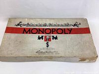 Top view of mostly white rectangular Monopoly box from 1930s featuring red stripe and black Monopoly text and graphics with wear and stains visible