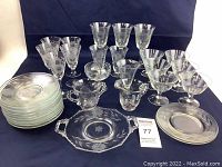 Front view of assorted glassware and plates arranged on a dark cloth background, showing plates, stemware, handled cups and tray.
