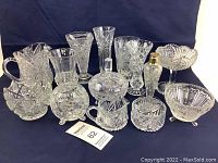 Full view of 13 clear cut and pressed glass pieces including vases, bowls, covered dish, and shaker on a dark background