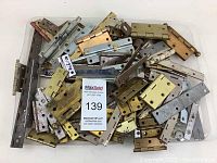 Top-down view of mixed assorted hinges including brass and chrome finishes, varying sizes and types scattered on a white surface.