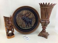 Clear frontal image showing three items: carved wooden figure, round moose wall plaque, and ornate metal vase.