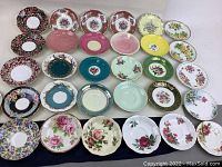 Overview photo showing 30 assorted fine china tea saucers laid out in rows with various colors and floral patterns, many with gold trim.
