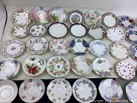 Top-down view of 40 assorted fine china tea saucers arranged in several rows showing floral patterns, different shapes and decorative borders.