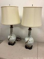 Pair of vintage decorative table lamps with cream-colored fabric shades and ceramic bases decorated with painted willow tree design.