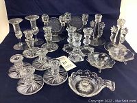 Photo showing the entire collection of 17 clear glass candle holders arranged on a dark cloth background. Shows variety in size, style, and design including starburst, fluted, ribbed, and etched motif holders.