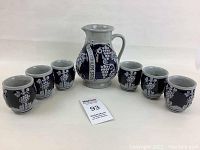 Photo showing full set of 7 pieces: 1 pitcher and 6 matching steins arranged in a semicircle with seller tag in front. Dark blue with white grapevine decoration, no visible chips.