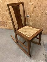 Side view of vintage wooden rocking chair with angled backrest and caned seat, rocker legs visible.