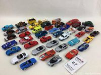 Photo shows a large variety of vintage diecast toy cars and trucks in various colors including blue, red, silver, yellow, green, and white. Cars include sports cars, pickups, convertibles, and utility trucks, arranged in multiple rows.