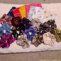 Wide view showing 5 assorted bandanas in navy blue, red, yellow, pink, white, and 13 assorted velvet and fabric scrunchies in multiple colors arranged on a white textured fabric.