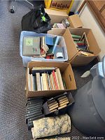 Overview of boxes and plastic tub containing books, CDs, and black rolling bag with zipper pockets.