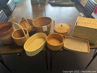 Top-down view of nine woven baskets arranged on a dark colored table, showcasing variety of shapes and sizes for general lot overview.